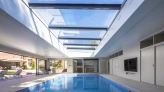 Intérieur d'une piscine moderne avec un plafond vitré, baignée de lumière naturelle. Des chaises longues sont disposées sur le côté et des portes coulissantes mènent à l'extérieur.