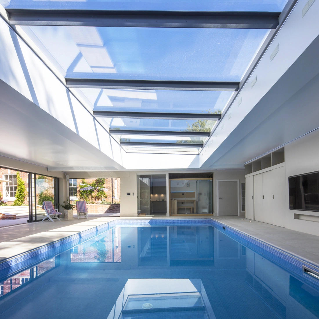 A spacious indoor pool area with a bright skylight, featuring a clear blue pool and modern minimalist design. Sliding doors lead to an outdoor space with lounge chairs.