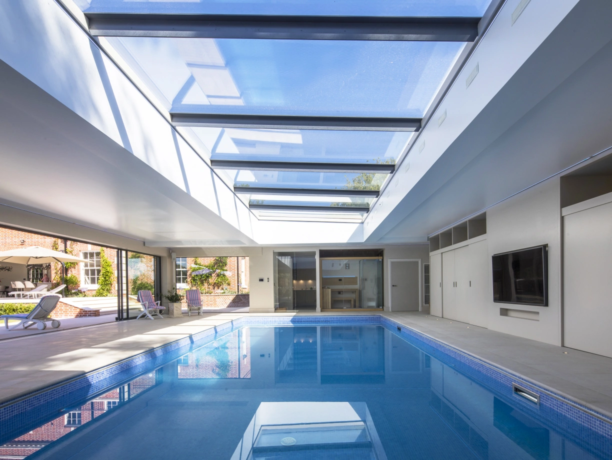 A spacious indoor pool area with a bright skylight, featuring a clear blue pool and modern minimalist design. Sliding doors lead to an outdoor space with lounge chairs.