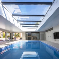 A modern indoor swimming pool with a glass ceiling, featuring sleek white walls and a flat-screen TV mounted on the wall. Sunlight streams through the skylights, illuminating the water. Comfortable lounge chairs are arranged around the pool area.
