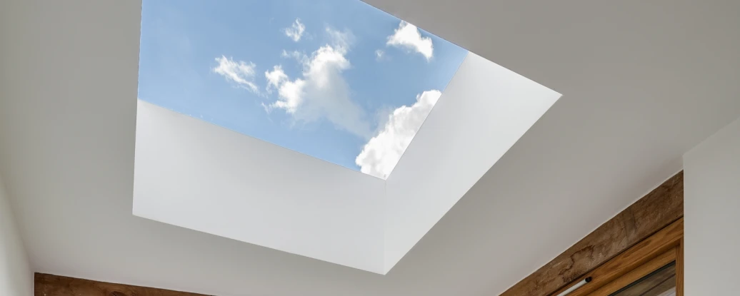 A cozy corner of a modern living space featuring a geometric patterned rug, plush cushions, and large windows offering a view of green fields and a blue sky with clouds. A skylight above lets in natural light, enhancing the inviting atmosphere.