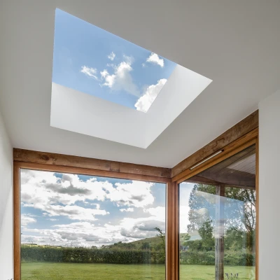 A cozy corner of a modern living space featuring a geometric patterned rug, plush cushions, and large windows offering a view of green fields and a blue sky with clouds. A skylight above lets in natural light, enhancing the inviting atmosphere.