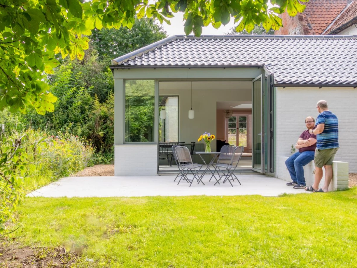 A modern home with a glass-walled extension opens to a lawn area, where two men are seated on a patio. Sunflowers are visible on the table, and lush greenery surrounds the property.