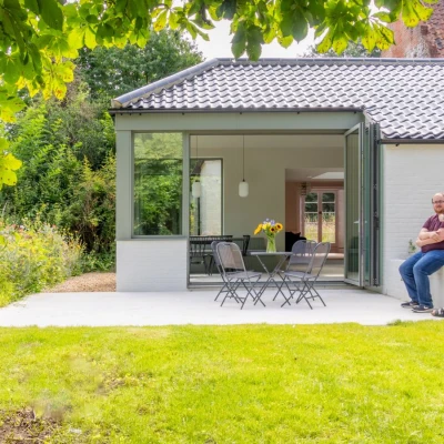 A modern home with a glass-walled extension opens to a lawn area, where two men are seated on a patio. Sunflowers are visible on the table, and lush greenery surrounds the property.