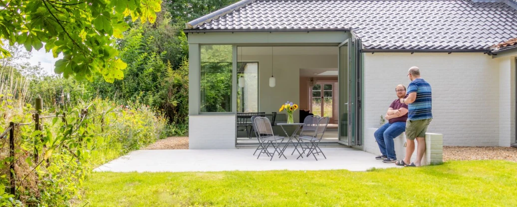 A modern home with a glass-walled extension opens to a lawn area, where two men are seated on a patio. Sunflowers are visible on the table, and lush greenery surrounds the property.