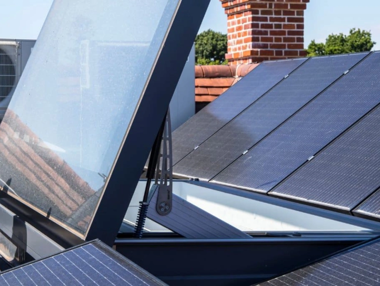 Paneles solares en un techo, con una estructura que parece abrirse y un fondo de cielo azul. Se observa un sistema de refrigeración al fondo y ladrillos rojos en el edificio.