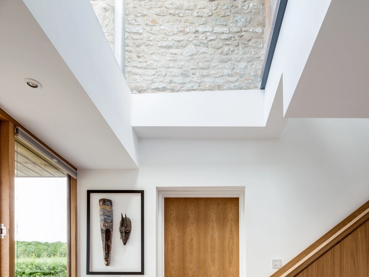 A modern interior hallway features a wooden door, a decorative mask on the wall, and a staircase leading upwards. Large windows allow natural light to flood the space, highlighting the smooth concrete floor and stone wall.