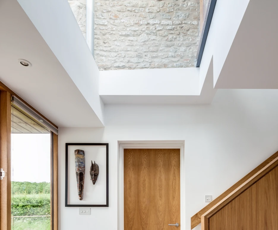 A modern interior hallway features a wooden door, a decorative mask on the wall, and a staircase leading upwards. Large windows allow natural light to flood the space, highlighting the smooth concrete floor and stone wall.