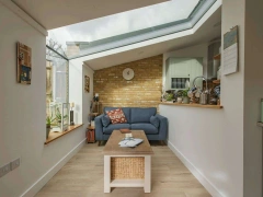 A cozy interior with a blue couch, a wooden coffee table, and a woven storage basket. Natural light floods the room through a slanted glass roof. A brick accent wall adds warmth, while plants and decor enhance the inviting atmosphere.