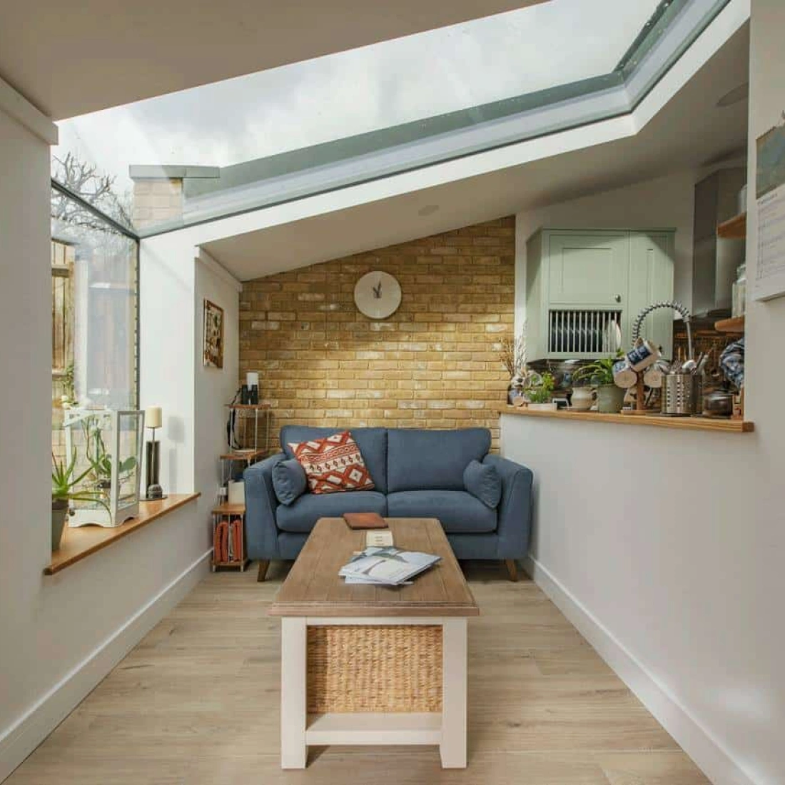 A cozy interior with a blue couch, a wooden coffee table, and a woven storage basket. Natural light floods the room through a slanted glass roof. A brick accent wall adds warmth, while plants and decor enhance the inviting atmosphere.