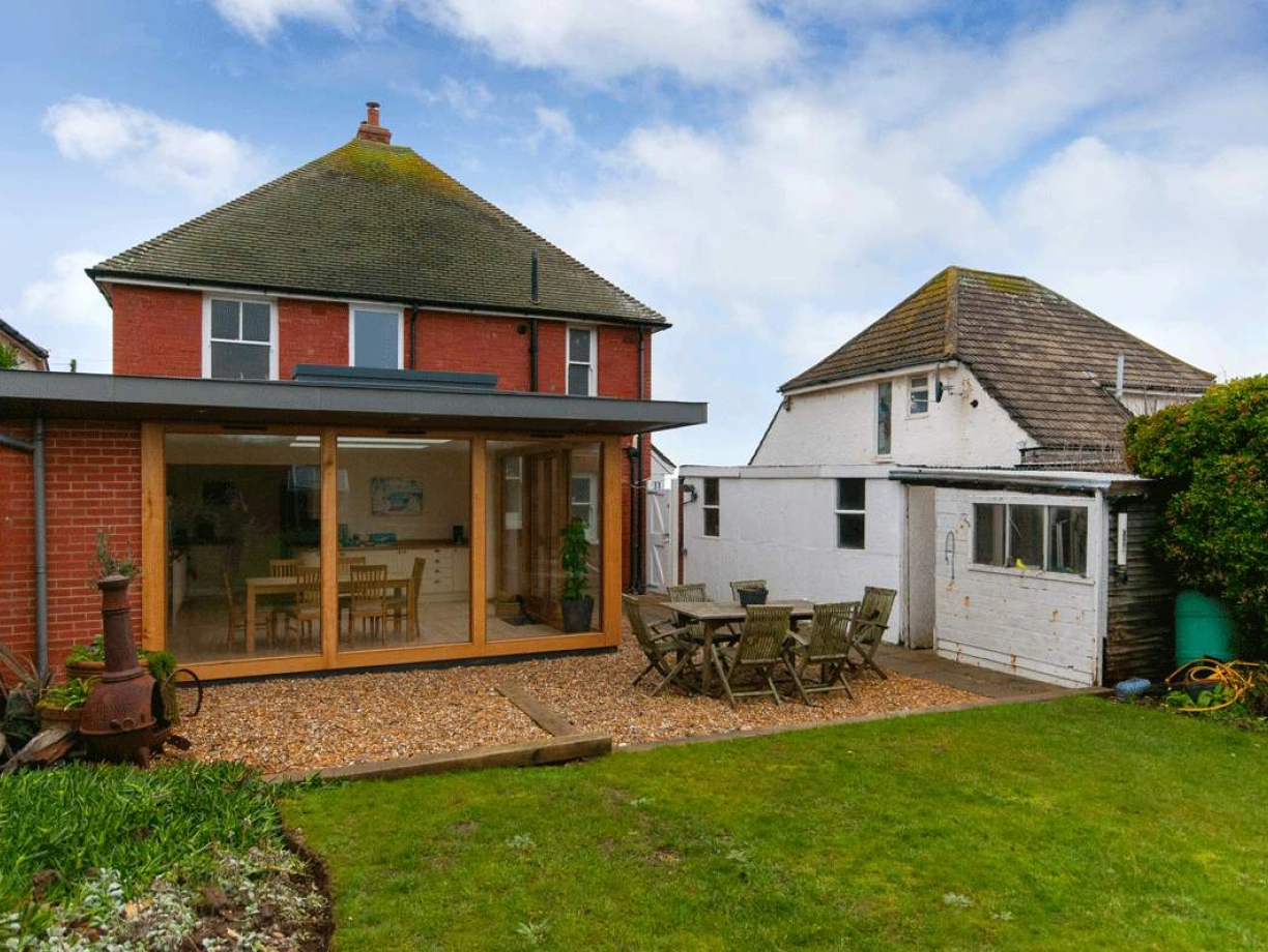 A modern two-story house with a large glass extension facing a well-maintained garden. There is a wooden dining table and chairs set up on a stone patio, surrounded by greenery. A smaller building is visible on the right, partially hidden by plants. The sky is partly cloudy.