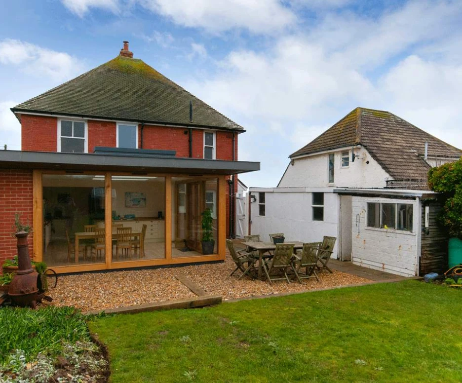 A modern two-story house with a large glass extension facing a well-maintained garden. There is a wooden dining table and chairs set up on a stone patio, surrounded by greenery. A smaller building is visible on the right, partially hidden by plants. The sky is partly cloudy.