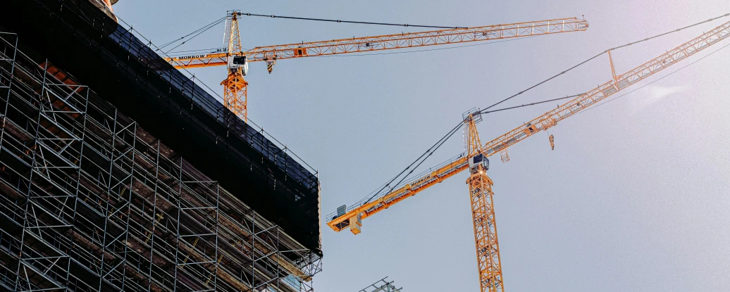 Two large construction cranes tower over a building site, with scaffolding wrapped around a structure. The bright sky appears in the background, reflecting sunlight.