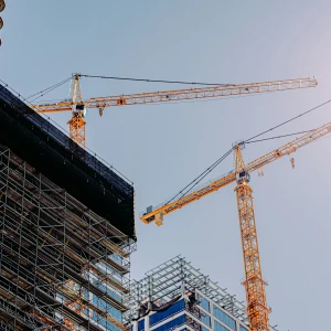 Two large construction cranes tower over a building site, with scaffolding wrapped around a structure. The bright sky appears in the background, reflecting sunlight.