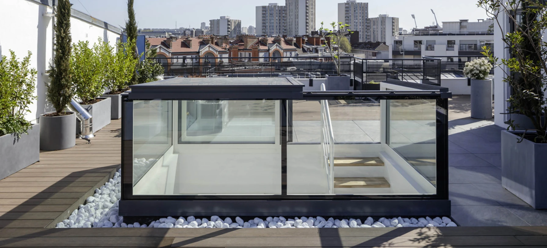 Toit-terrasse moderne avec une structure en verre et des plantes, offrant une vue sur la ville. Des galets blancs entourent la structure, et des immeubles se dressent en arrière-plan sous un ciel clair.