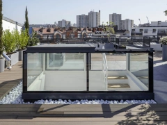 A modern rooftop terrace featuring a glass-walled structure surrounded by greenery and white stones. The skyline shows residential buildings in the background under a clear blue sky.