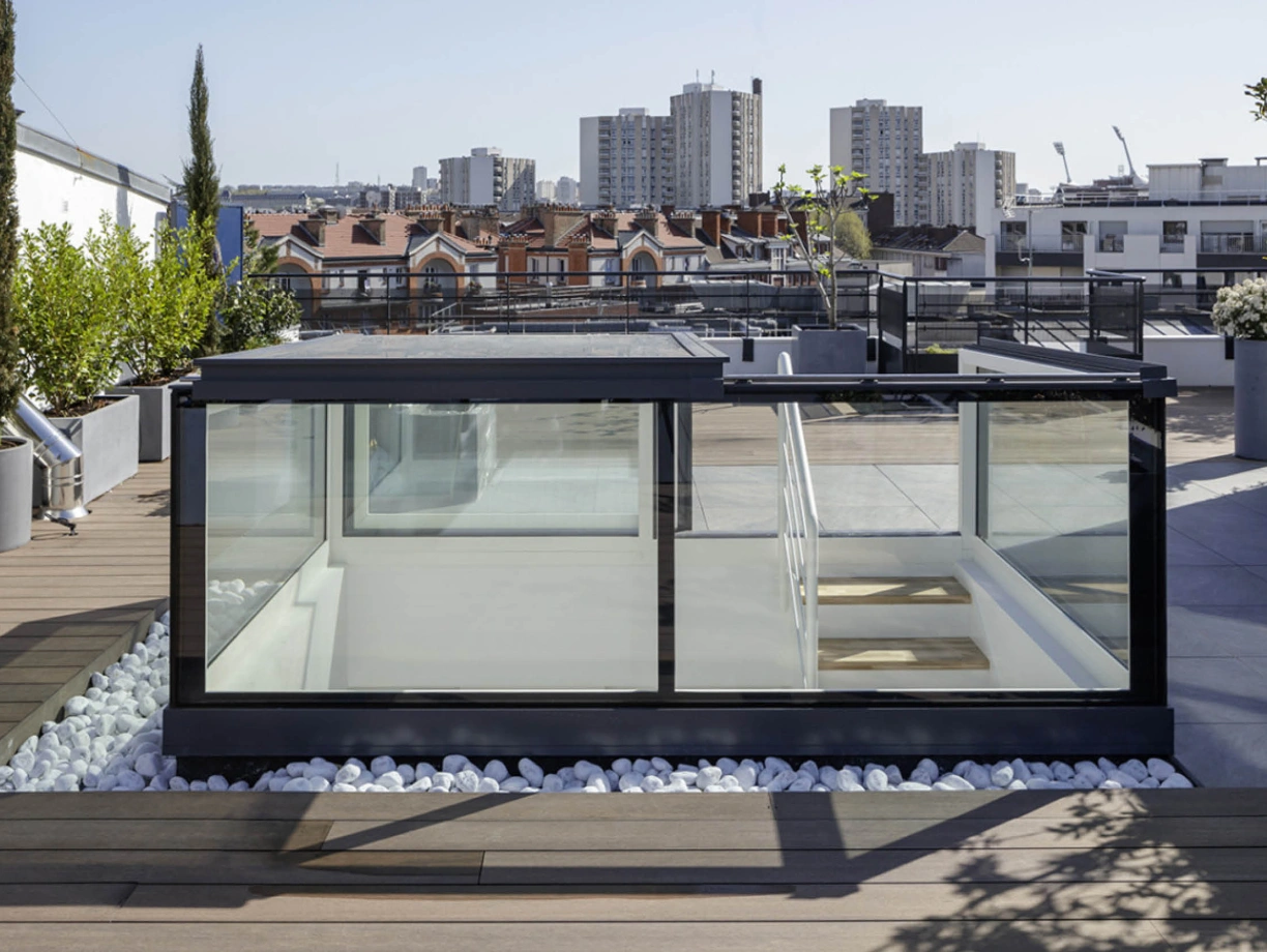 A modern rooftop terrace featuring a glass-walled structure surrounded by greenery and white stones. The skyline shows residential buildings in the background under a clear blue sky.
