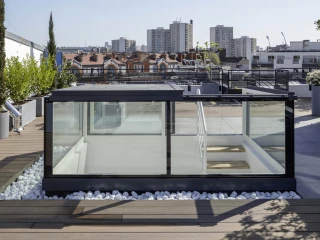 Toit-terrasse moderne avec une structure en verre et des plantes, offrant une vue sur la ville. Des galets blancs entourent la structure, et des immeubles se dressent en arrière-plan sous un ciel clair.