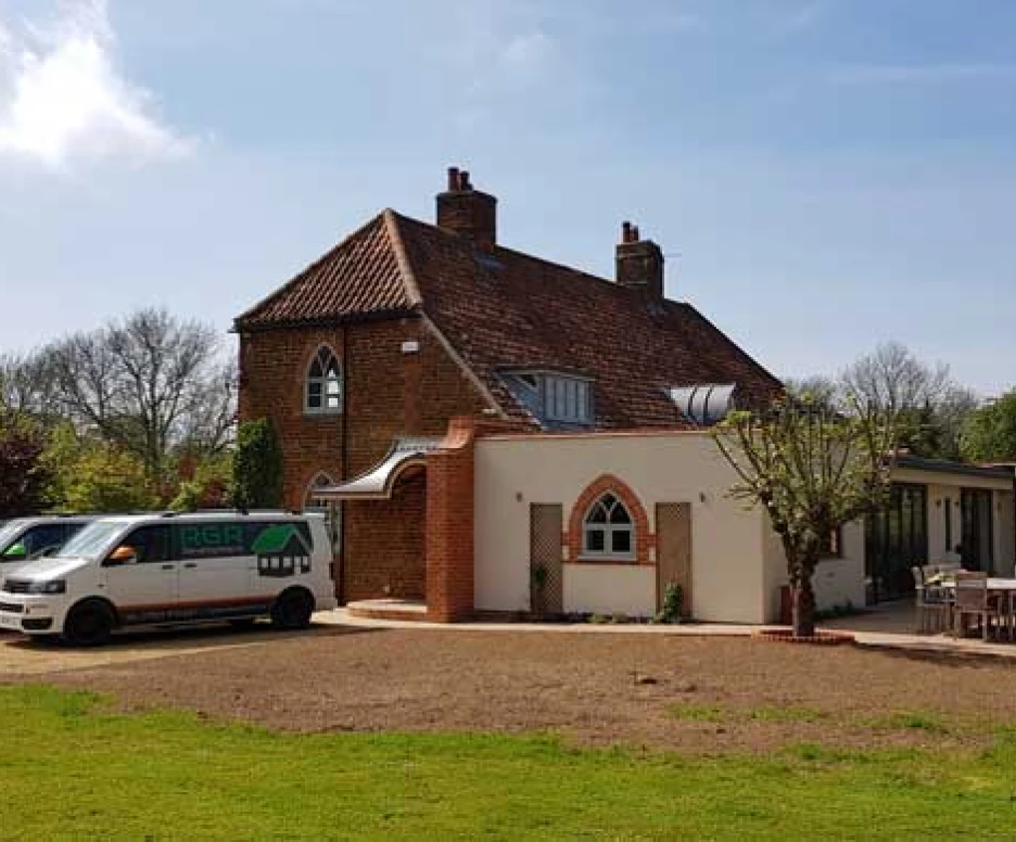 A spacious, modern house with a mix of traditional and contemporary architectural features. Several vehicles are parked in the driveway, and a lawn surrounds the property with trees and greenery in the background.