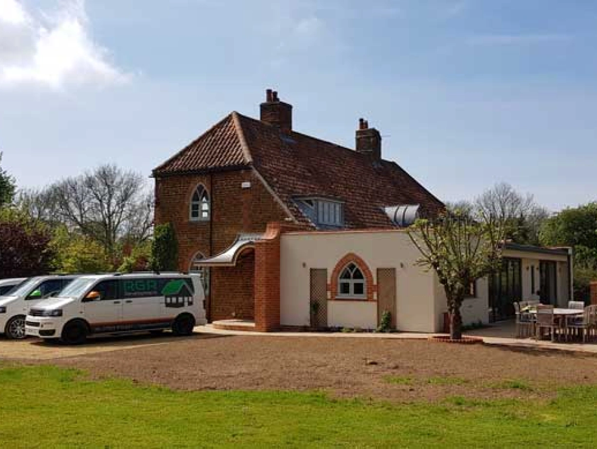 A spacious, modern house with a mix of traditional and contemporary architectural features. Several vehicles are parked in the driveway, and a lawn surrounds the property with trees and greenery in the background.