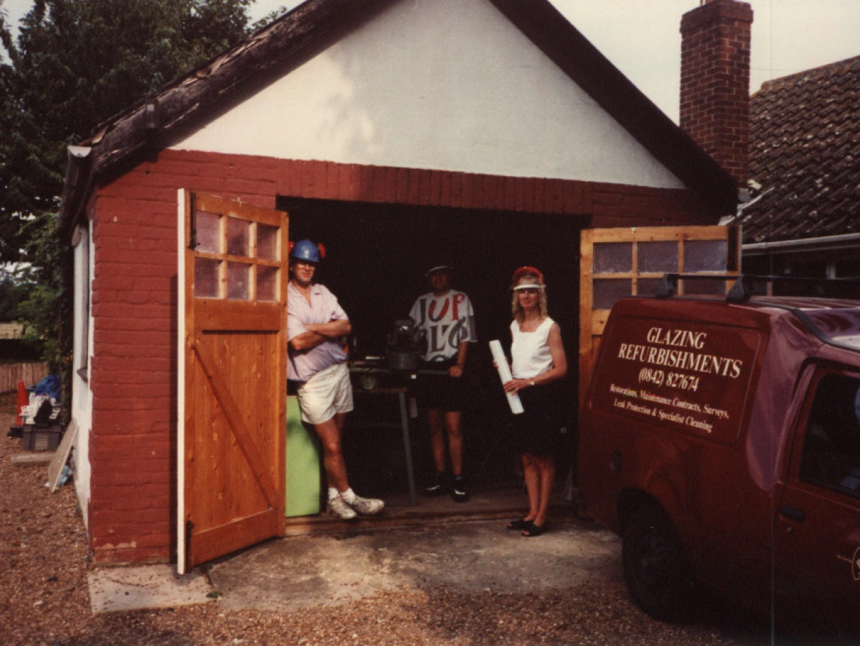 A man wearing a helmet and casual clothing leans against a brick building, while a woman in a hat and a t-shirt stands beside him. Another person is visible inside the garage, which features a small work area. A red van with "Glazing Refreshments" written on it is parked nearby. The setting appears to be a rural area with trees in the background.