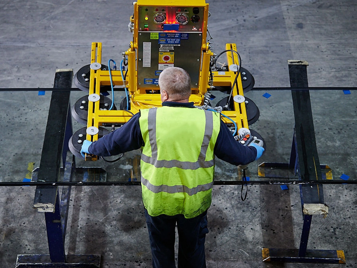 A worker in a yellow safety vest operates a glass handling machine, guiding a large sheet of glass in a warehouse setting. The machine features suction cups and control panels.