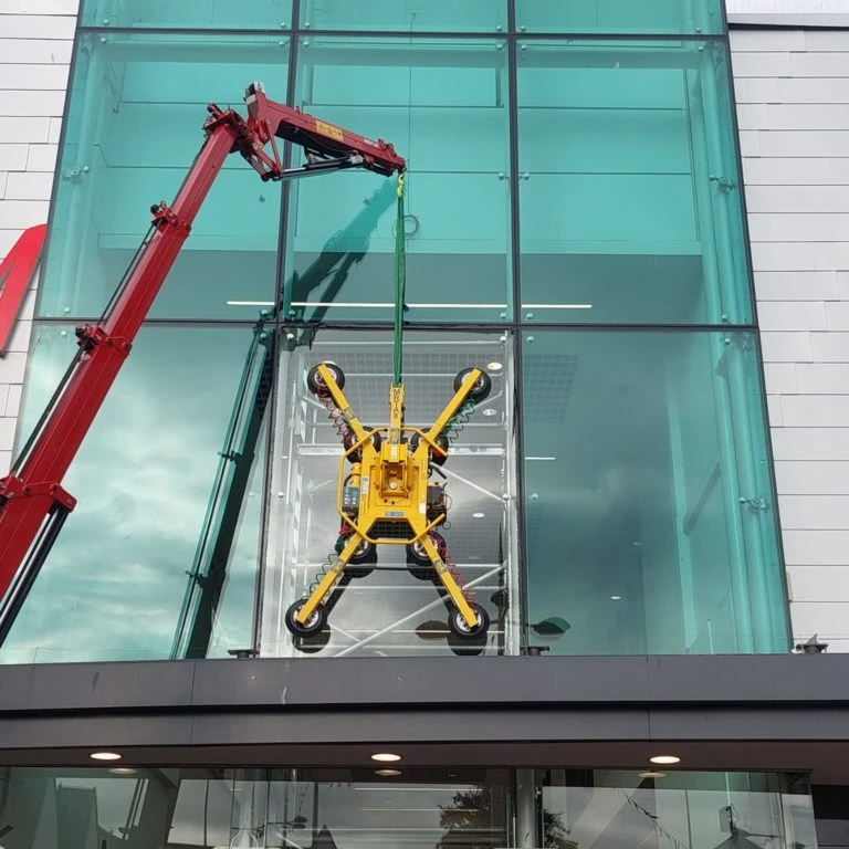 A yellow robotic glass lifter is suspended from a red crane, positioned in front of a large glass facade. The lifter is attaching to a large glass panel while a cloudy sky is visible in the background.