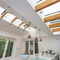 Luminoso cucina con soffitto alto, dotato di finestre a tetto in vetro e travi in legno. Eleganti lampade pendenti d'oro illuminano il piano di lavoro chiaro. Una grande finestra a vetro si affaccia su un giardino verde.