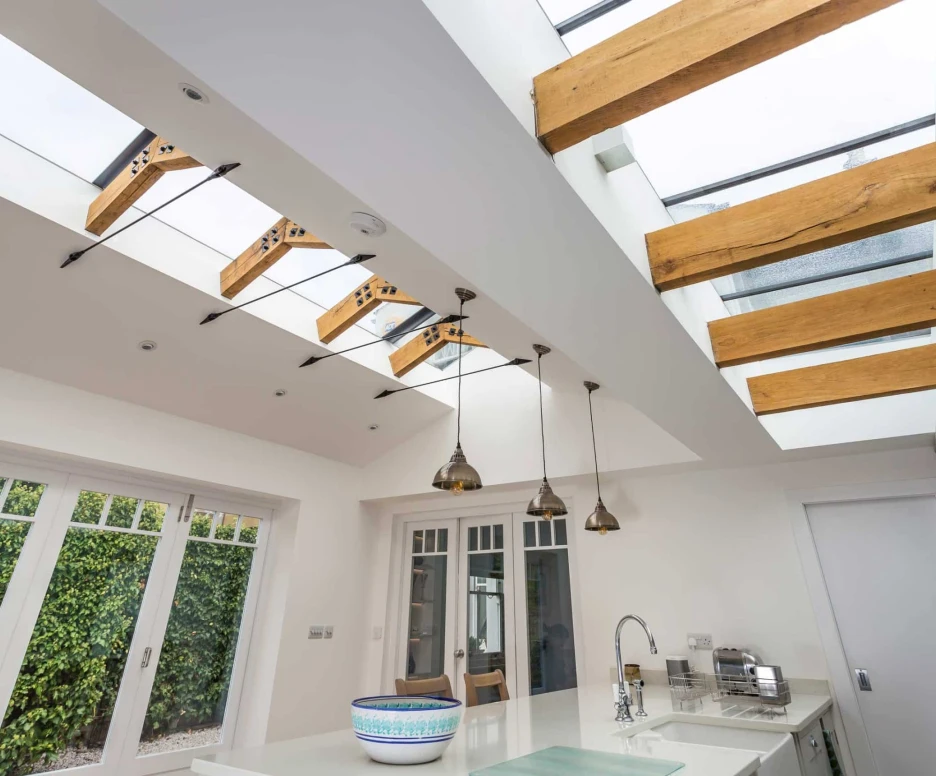 Modern kitchen featuring a skylight with wooden beams. The space is brightly lit and includes pendant lighting, white cabinetry, and a large central island with a decorative bowl. Greenery is visible through large windows.