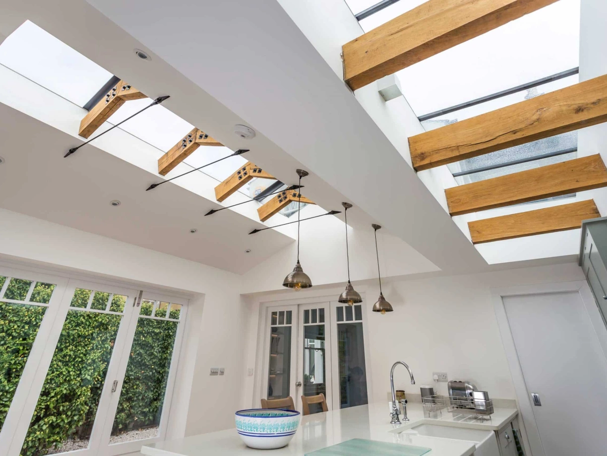 Modern kitchen featuring a skylight with wooden beams. The space is brightly lit and includes pendant lighting, white cabinetry, and a large central island with a decorative bowl. Greenery is visible through large windows.