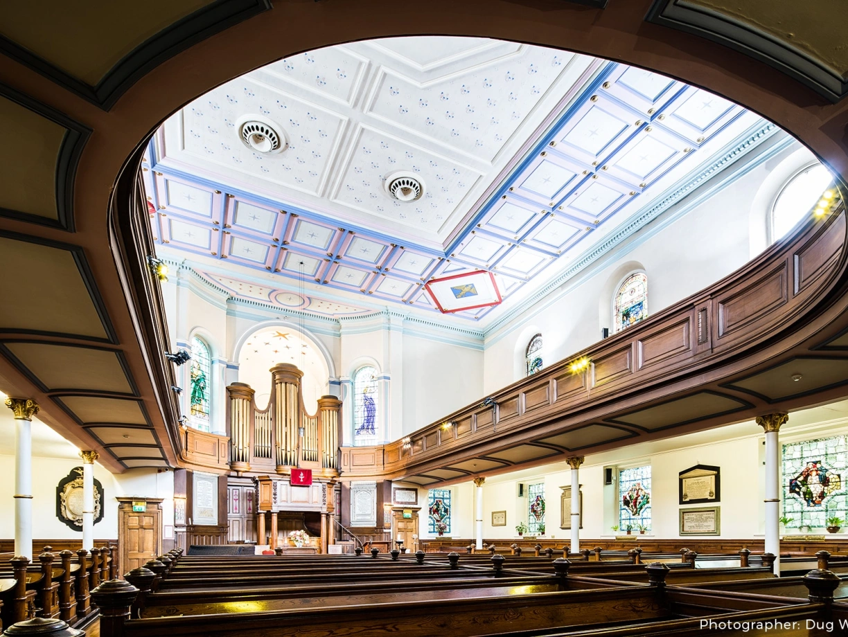 Interior view of a church featuring wooden pews, ornate stained glass windows, and a decorative ceiling with intricate designs. A prominent pipe organ is visible at the front, adding to the serene atmosphere.