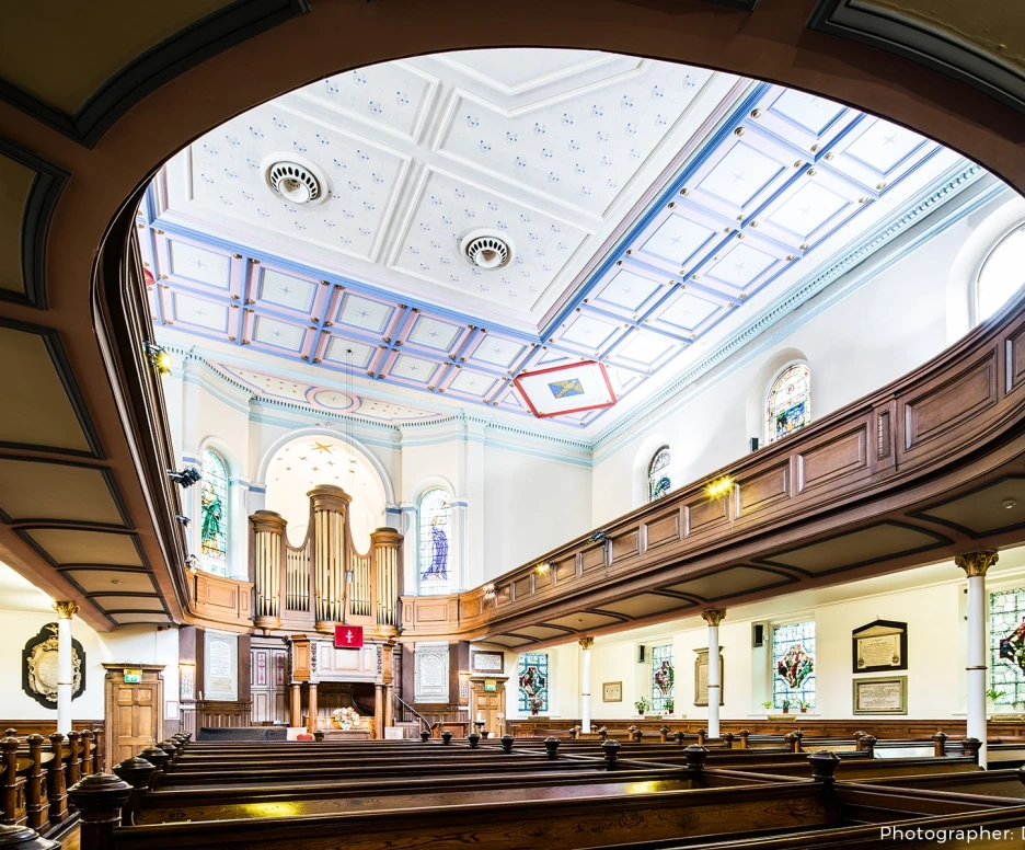 Interior view of a church featuring wooden pews, ornate stained glass windows, and a decorative ceiling with intricate designs. A prominent pipe organ is visible at the front, adding to the serene atmosphere.