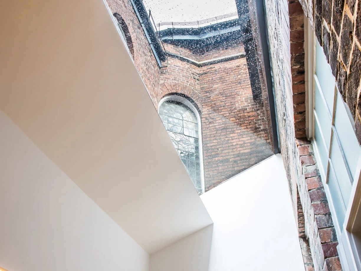 A modern interior with a skylight allowing rain to fall inside. Brick walls contrast with a minimalist white ceiling. A glimpse of an arched window is visible outside.