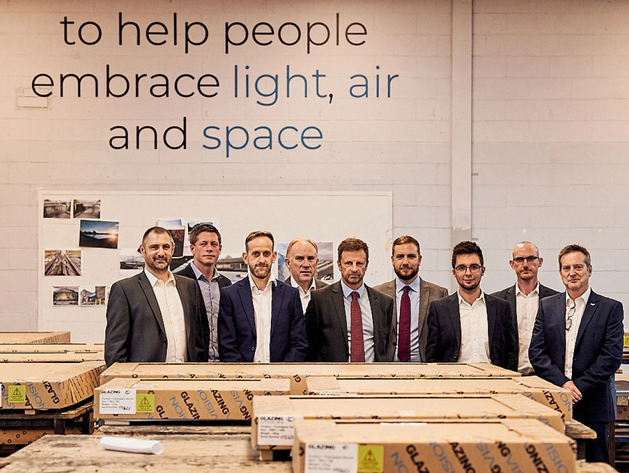 Eine Gruppe von neun Männern in Anzügen steht in einem Raum mit Holzkisten. Im Hintergrund ist ein Wandtext zu sehen, der "to help people embrace light, air and space" sagt. Alle schauen in die Kamera und die Stimmung ist freundlich.