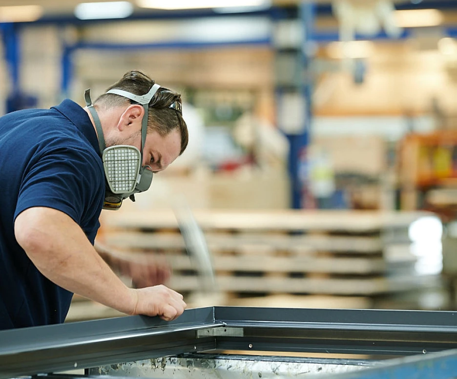 A worker in a blue shirt and mask is closely inspecting and adjusting a metal frame in a workshop filled with various equipment and materials.