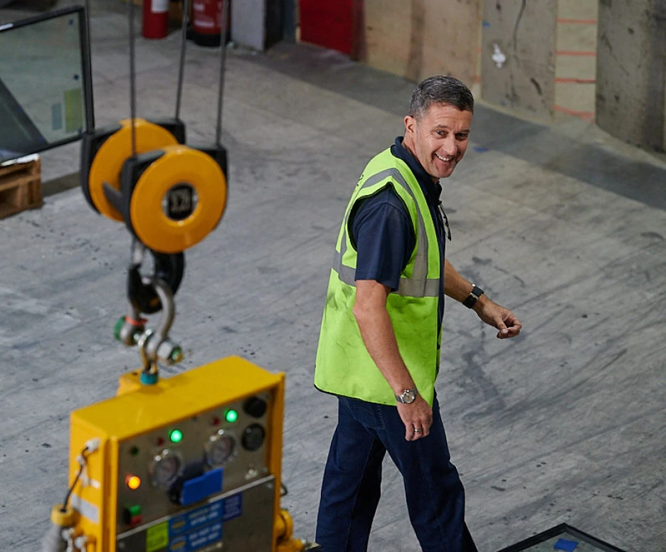 A man in a green safety vest walks through a warehouse, smiling, with a glass panel and industrial equipment in the background.