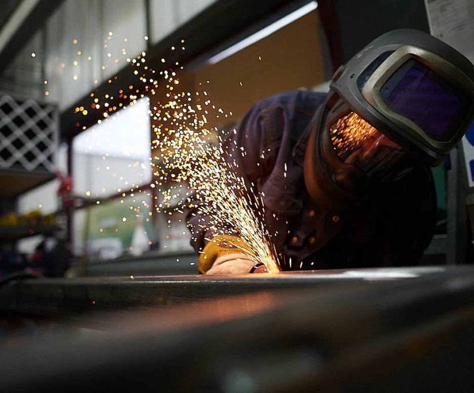 Un soudeur travaille sur un métal, produisant des étincelles brillantes. Il porte un casque de protection et des lunettes de sécurité, concentré sur sa tâche dans un atelier.