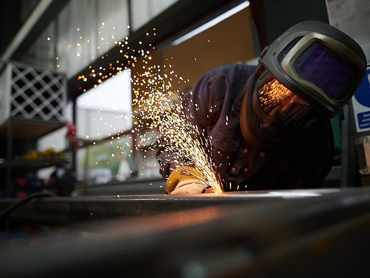 Un soldador trabaja en un taller, creando chispas brillantes mientras utiliza una herramienta de corte. La luz refleja un ambiente industrial.