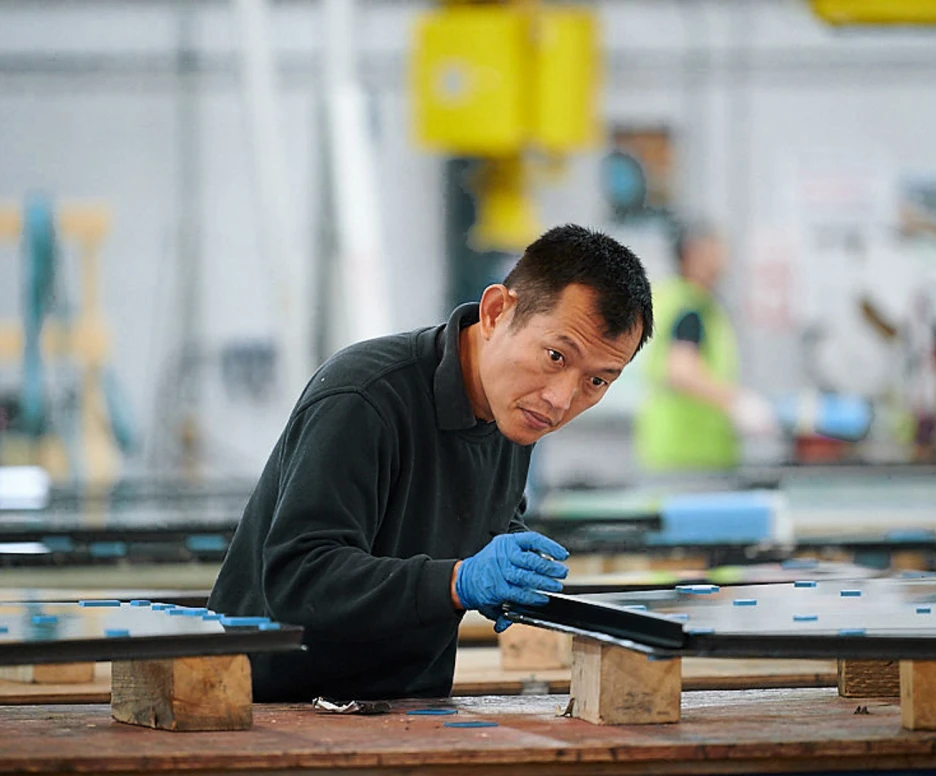 Un ouvrier travaille sur des panneaux de verre dans une usine. Il porte des gants bleus et se concentre sur sa tâche, tandis que d'autres personnes sont en arrière-plan.