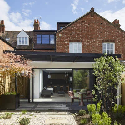 Modern garden space featuring a sleek glass extension with a wooden deck, surrounded by greenery and a mix of plants. The backdrop includes traditional brick houses, with a clear blue sky above.