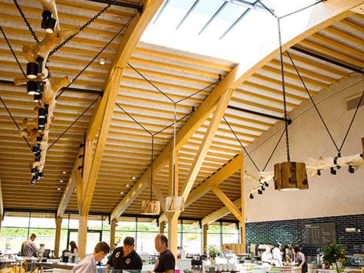 A spacious modern café with high wooden beams and a skylight. Staff members prepare food in a well-organized area featuring glass display cases filled with fresh dishes. Natural light illuminates the interior, creating a welcoming atmosphere.