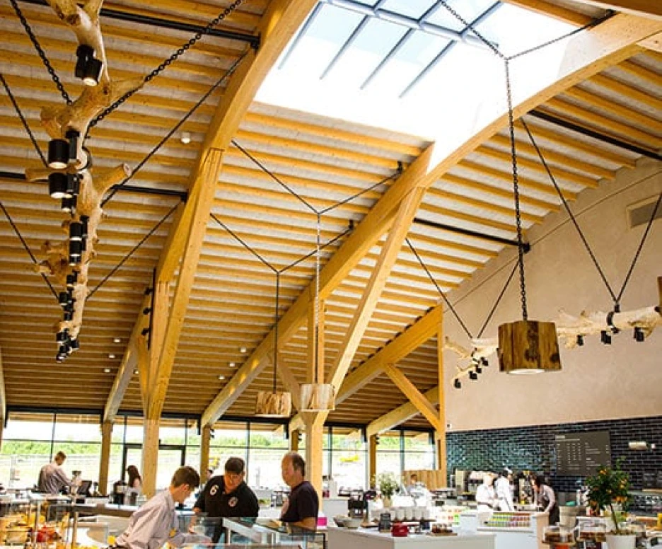 A spacious modern café with high wooden beams and a skylight. Staff members prepare food in a well-organized area featuring glass display cases filled with fresh dishes. Natural light illuminates the interior, creating a welcoming atmosphere.