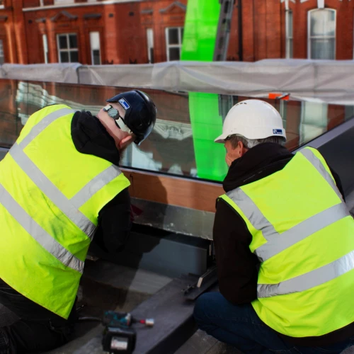 Two construction workers, wearing safety vests and helmets, are kneeling and working on a reflective metal surface. They are on a rooftop, with a green tarp visible in the background and red brick buildings. Tools are scattered around them.