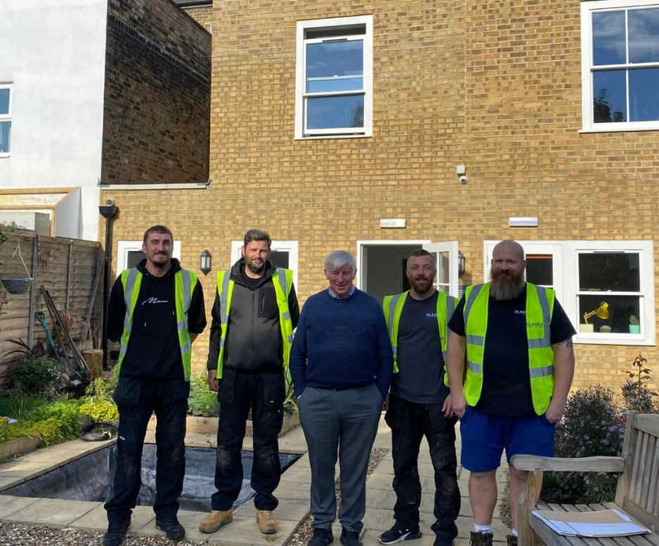 Five men stand together in front of a house with a brick facade. All wear high-visibility vests. The garden area features flowers, a bench, and a partially dug area. Sunlight brightens the scene.