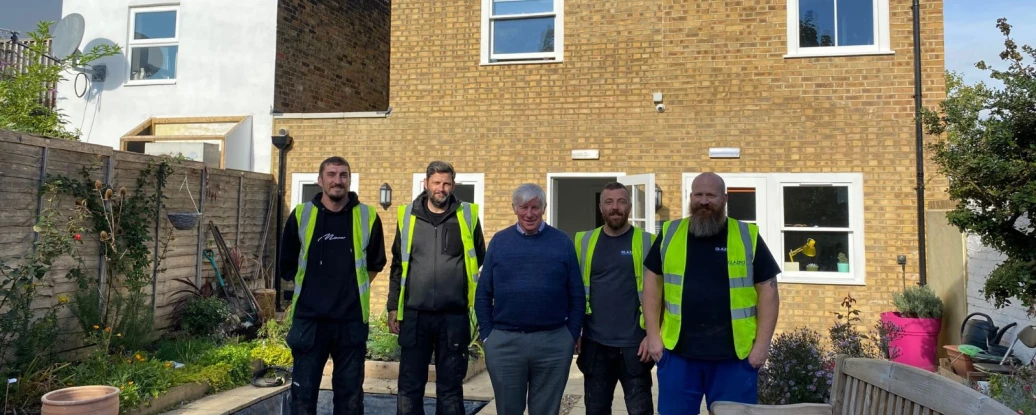 Five men stand together in front of a house with a brick facade. All wear high-visibility vests. The garden area features flowers, a bench, and a partially dug area. Sunlight brightens the scene.