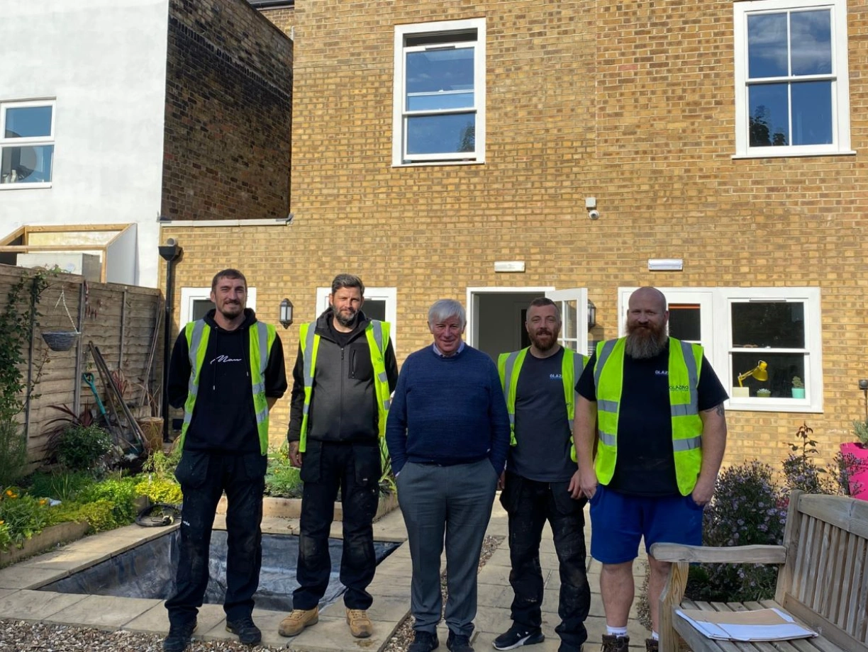 Five men stand together in front of a house with a brick facade. All wear high-visibility vests. The garden area features flowers, a bench, and a partially dug area. Sunlight brightens the scene.