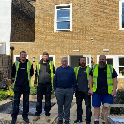 Five men stand together in front of a house with a brick facade. All wear high-visibility vests. The garden area features flowers, a bench, and a partially dug area. Sunlight brightens the scene.