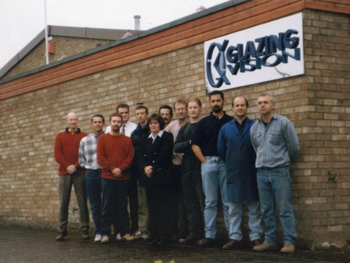 A group of twelve individuals poses in front of a brick wall featuring a sign that reads "Glazing Vision." The group includes men and one woman, all dressed in casual clothing. The setting appears to be an outdoor location near a building.