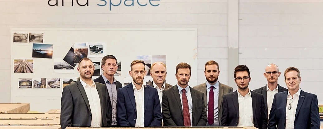 A group of nine professional men stands in a warehouse, posing in front of stacked wooden crates. A wall behind them features a slogan about embracing light, air, and space.