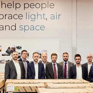 A group of nine professional men stands in a warehouse, posing in front of stacked wooden crates. A wall behind them features a slogan about embracing light, air, and space.